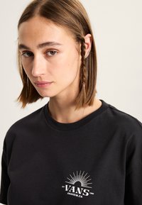 Camiseta de algodón negra con un estampado gráfico blanco de "VANS" con diseño de rayos de sol en el lado izquierdo del pecho. Cuello redondo casual y mangas cortas.