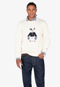 Suéter de punto crema con un diseño de perro azul y blanco con detalles bordados. Combinado con vaqueros de mezclilla oscuros. Se aprecia un cuello plano.