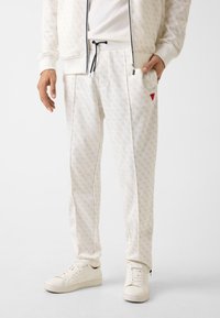 Weiße Jogginghosen mit dezentem Muster, verstellbarem schwarzen Kordelzug, Seitentaschen und rotem Akzentdreieck. Kombiniert mit weißen Turnschuhen.