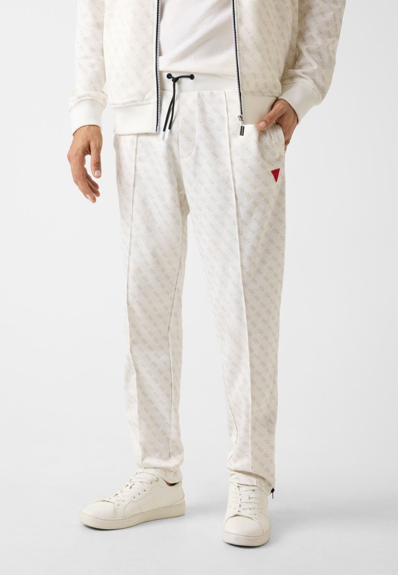 Witte joggingbroek met patroon en subtiel ontwerp, verstelbare zwarte trekkoord, zijzakken en een rood accent driehoek. Gecombineerd met witte sneakers.