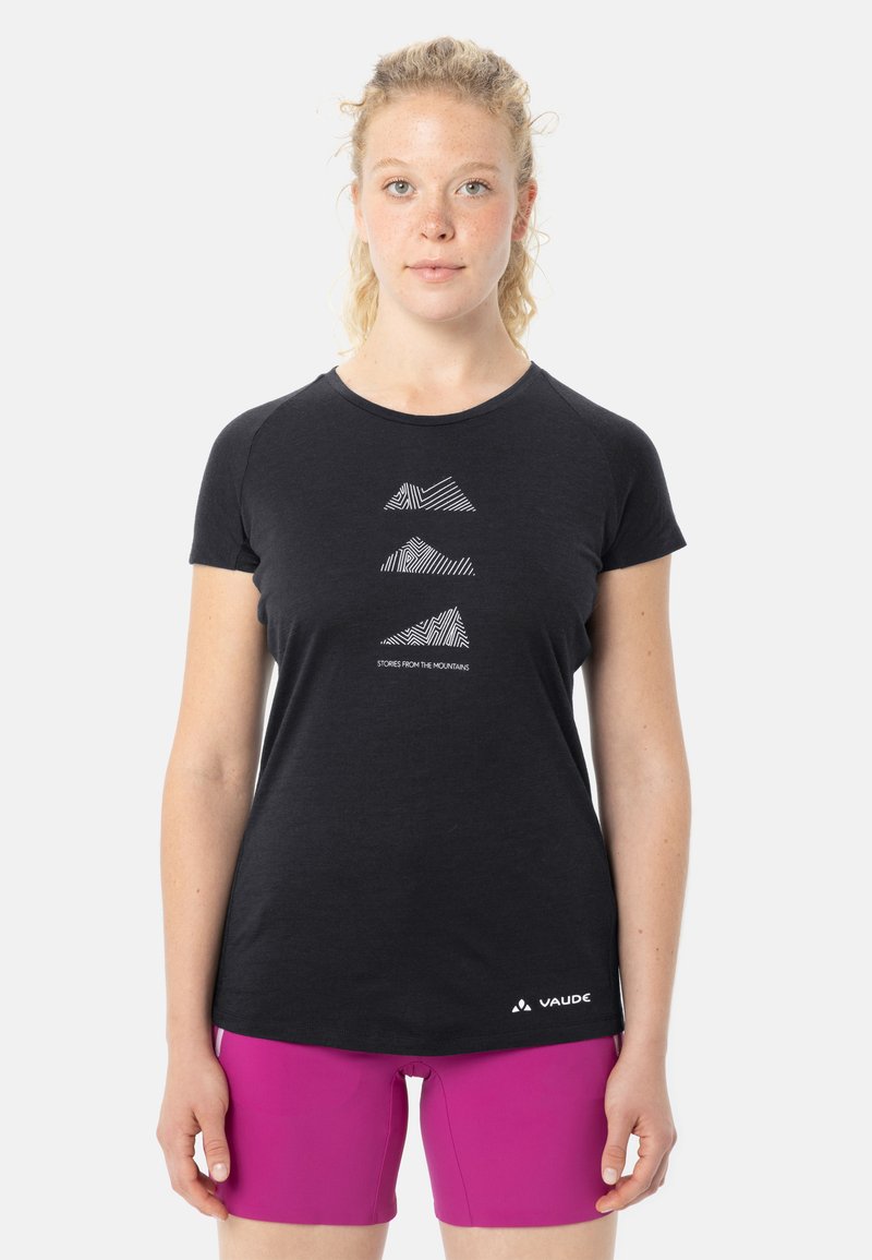 Persona bionda con capelli ricci indossa una maglietta nera con disegni di montagne e pantaloncini magenta, in piedi davanti a uno sfondo bianco.