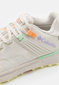 Ljust beige atletisk sko med nätmaterial, orange och gröna accenter, och strukturerad sula med ett zigzag mönster. Logotyp på sidan.