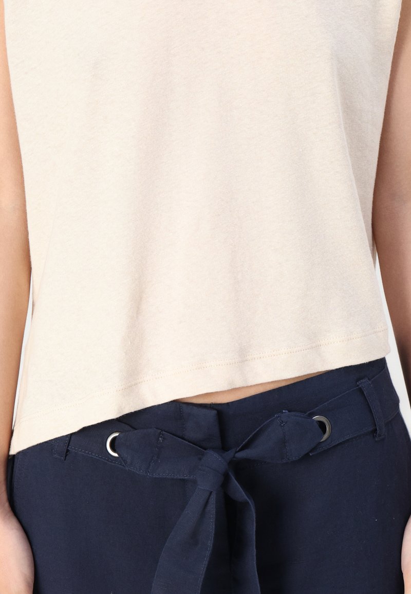 Beige Cropped-Top, kombiniert mit navyblauen Hosen, die einen Stoffgürtel haben, der in einer Schleife an der Taille gebunden ist.