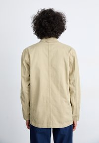 Beige cotton jacket with a straight cut, long sleeves, and a back seam detail, paired with dark blue trousers.