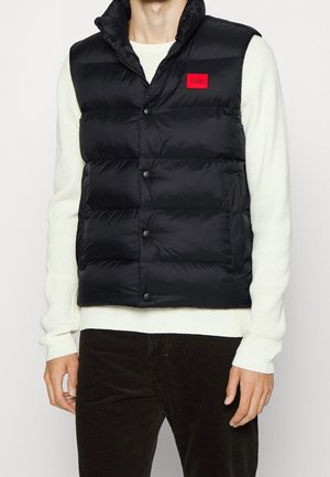 Mann trägt eine schwarze gesteppte Weste mit rotem Hugo-Logo auf der Brust über einem weißen Strickpullover und dunkler Cordhose.