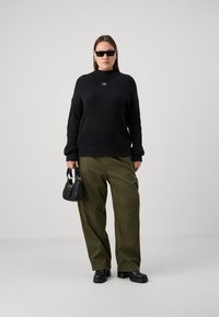 Pull noir à côtes avec col montant, pantalon vert à jambes larges, sac à main noir et lunettes de soleil oversize noires. Chaussures à talons épais noires.