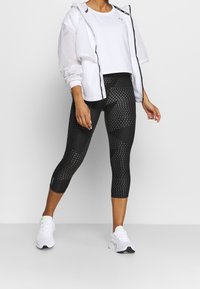 Leggings negros con un diseño geométrico, combinados con una camiseta corta blanca y una chaqueta ligera blanca. Zapatillas deportivas blancas.
