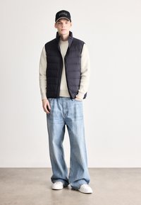 Melna pūkaina veste virs krēmkrāsas ribota džempera, kombinēta ar vaļīgiem gaismas zilas krāsas džinsa biksēm un baltām sporta kurpēm; papildināta ar melnu cepuri.