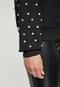 Felpa nera con maniche decorate, caratterizzata da perle bianche e strass lucenti. Tessuto liscio e design con polsini attillati.