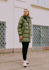 Ieliekamais olīvzaļais mētelis ar rāvējslēdzēju priekšā, kapuci un kontrastējošiem aukliņām, uzvilkts virs pelēka augsta kakla krekla un melniem biksēm, ar baltām sporta kedām.