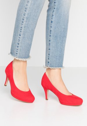 Ben klædt i lyseblå jeans med fransk kant og klare røde suedepumps, og står på et hvidt gulv.