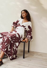 Langes, bedrucktes Kleid in Weiß mit maroonfarbenen botanischen Mustern. Es verfügt über weite Ärmel, einen tiefen V-Ausschnitt und eine fließende Silhouette.