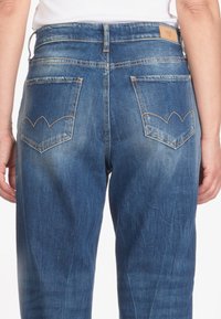 Pantalones vaqueros de denim azul con una cintura baja, dos bolsillos traseros que presentan un distintivo patrón de costura en forma de chevron y un ligero desgaste.