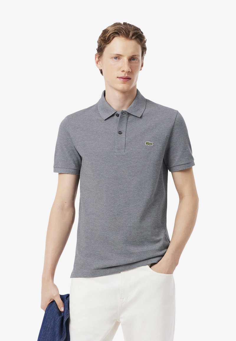 Graues Poloshirt aus Baumwolle mit klassischem Kragen und kurzen Ärmeln. Lacoste-Logo in Grün auf der Brust, kombiniert mit weißer Hose.