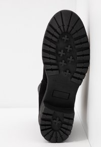 Semelle de botte noire avec des sculptures profondes et des motifs en forme de croix reposant contre un mur d’angle blanc.