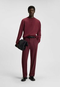 Bordeaux-rood langwerpig shirt met ronde hals en logo, gecombineerd met bijpassende op maat gemaakte broeken. Zwarte riem en schoenen. Houdt een zwart jack vast.