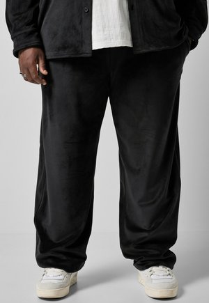 Persona que lleva pantalones negros holgados, zapatillas blancas, una camisa blanca acanalada y una chaqueta negra, con una mano en el bolsillo y la otra relajada.
