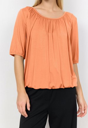 Peach short-sleeve blouse with a round neckline, gathered detailing at the shoulders, and a relaxed fit, paired with black trousers.