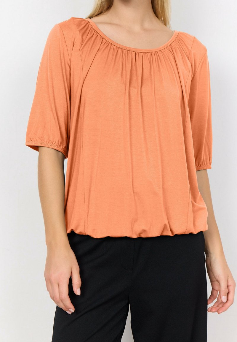 Blusa de manga corta melocotón con escote redondo, detalles fruncidos en los hombros y un corte relajado, combinada con pantalones negros.