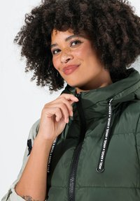 Gilet verde in piuma con zip frontale, cappuccio e lacci. Il tessuto ha una texture liscia con una finitura lucida e un accento di logo nero.