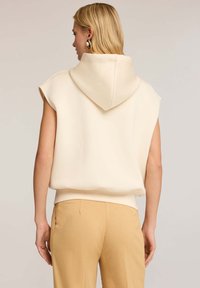 Sudadera sin mangas beige con capucha, hecha de material suave, presenta un ajuste relajado y dobladillo acanalado. Combinada con pantalones de color crema.