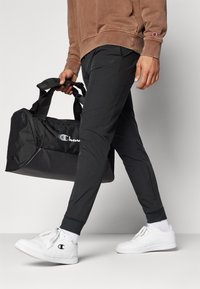 Borsa duffel nera con logo, indossata con pantaloni grigi a gamba stretta e sneaker bianche. I pantaloni hanno polsini elasticizzati; la borsa presenta doppi manici e chiusura con zip.