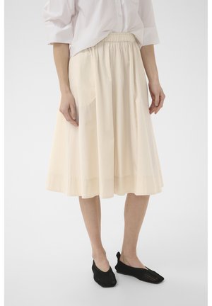 Femme portant une chemise blanche boutonnée, une jupe crème mi-longue froncée avec une taille élastique, et des chaussures noires tissées à enfiler, debout sur un sol blanc.