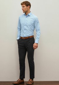 Helles, blaues Hemd mit strukturiertem Kragen, kombiniert mit dunklen Hosen sowie brauem Gürtel und Schuhen, vor einem neutralen Hintergrund stehend.