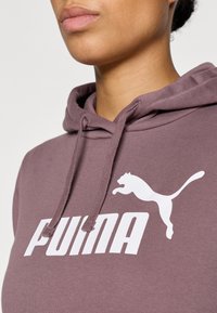 Fialová mikina s kapucňou a šnúrkou, s veľkým bielym logom "PUMA" a bielou skáčucou grafikou pumy. Mäkká, textúrovaná látka.