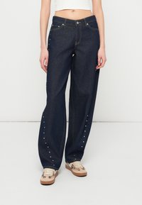 Σκούρα μπλε τζιν παντελόνια με άνετη εφαρμογή, με ασημένιες μεταλλικές λεπτομέρειες κατά μήκος των εξωτερικών ραφών και ανοιχτόχρωμες ραφές. Συνδυασμένα με μπεζ αθλητικά παπούτσια.