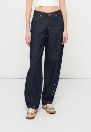 Jean azul escuro com um corte largo, apresentando tachas prateadas ao longo das costuras externas e costura clara. Combinado com ténis bege.