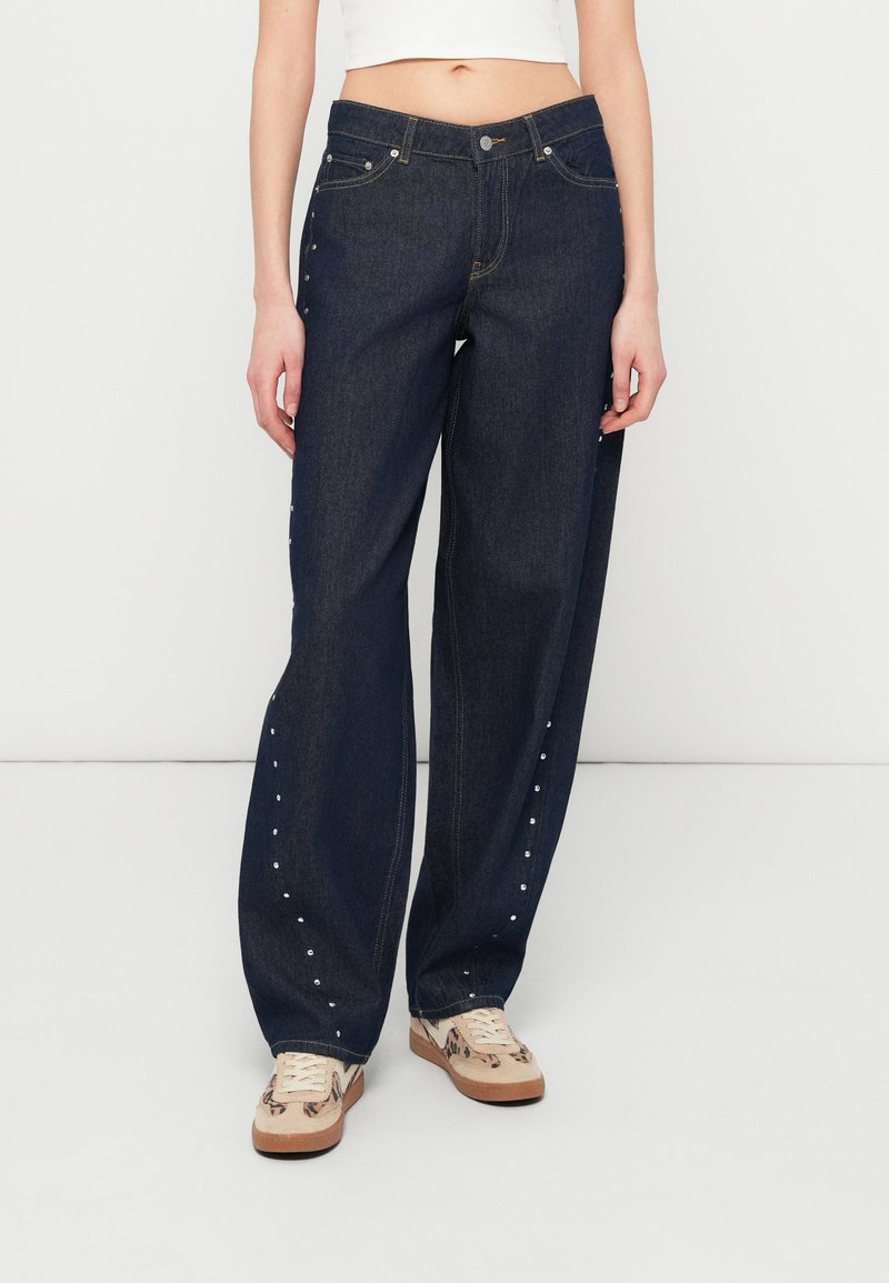 Σκούρα μπλε τζιν παντελόνια με άνετη εφαρμογή, με ασημένιες μεταλλικές λεπτομέρειες κατά μήκος των εξωτερικών ραφών και ανοιχτόχρωμες ραφές. Συνδυασμένα με μπεζ αθλητικά παπούτσια.