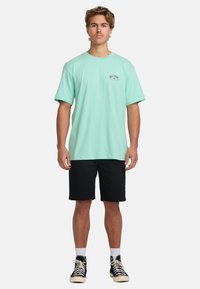 Mint green short-sleeve t-shirt with a round logo on the chest, paired with black shorts and black sneakers with white laces and ankle socks.