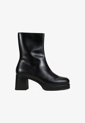 Botín negro de cuero hasta el tobillo con una textura suave y brillante, puntera redondeada y tacón cuadrado. Presenta un caña ajustada y elementos de diseño mínimos.