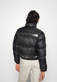 Jachetă puf din material negru, cropped, cu guler înalt, textură lucioasă și detaliu cu logo pe spate. Echipată cu mansete și tiv elastic.