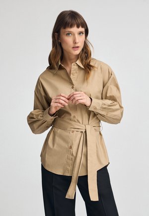 Camicia in cotone beige con colletto a bottoni, maniche lunghe e vita legata. Presenta una texture liscia e un tessuto morbido. Sono indossati pantaloni neri.