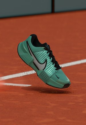 Chaussure de sport Nike verte et noire suspendue au-dessus d'un court de tennis en terre battue rouge près d'une ligne de délimitation blanche avec de la poussière d'argile visible.