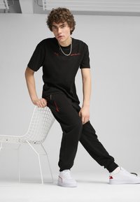 Czarna koszulka i dresowe spodnie, obie z czerwonymi akcentami. Koszulka ma logo, a model ma na sobie białe sneakersy. Tło jest neutralne, minimalistyczne.