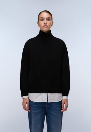 Maglione nero a collo alto con una vestibilità morbida, realizzato in un morbido materiale a maglia, abbinato a una camicia bianca e grigia a righe sotto.
