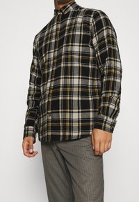 Camisa de franela a cuadros en negro, gris y amarillo con cuello abotonado y mangas largas, que presenta un ajuste relajado y una tela texturizada.