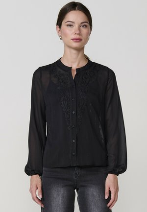 Blusa negra transparente con mangas largas, botón en el frente, detalles bordados a lo largo del escote y puños fruncidos. Textura suave y ligera.