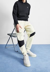 Schwarzer Kapuzenpullover mit Logo, kombiniert mit hellbeigen Jogginghosen mit schwarzen Akzenten und weißen Sneakers auf einem blauen Teppich.