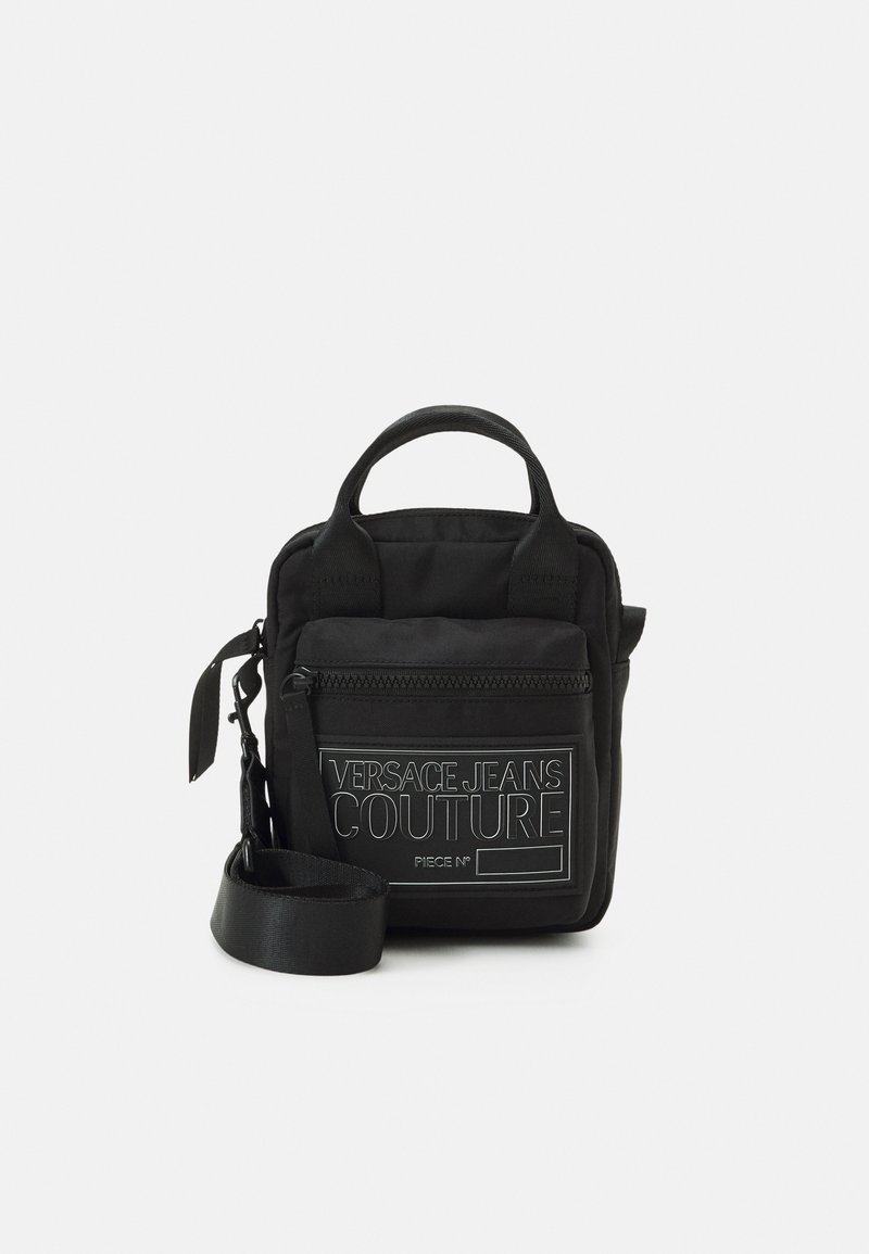 Černá textilní taška s krátkými madly, nastavitelným popruhem, přední kapsou na zip a kontrastním bílým detailem s logem značky. Kompaktní obdélníkový tvar.