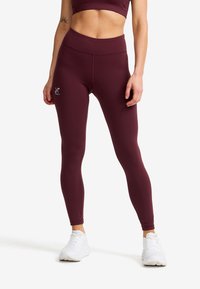 Kvinna iklädd högmidjade, mörklila leggings och en matchande sport-bh, stående med vita träningsskor på en vit bakgrund.