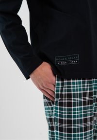 Czarna koszula z długim rękawem z zielono-białą metką, w połączeniu z zielono-czarnymi spodnimi w kratę z białymi akcentami i kieszenią.