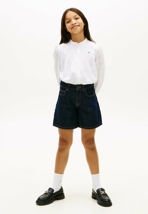 Ragazza in piedi con le mani dietro la schiena, indossa una camicia bianca con bottoni, shorts in denim scuro, calzini bianchi e mocassini neri su sfondo semplice.