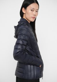 Σκούρο ναυτικό μπουφάν με οριζόντιες ραφές, εφαρμοστή σιλουέτα, αποσπώμενη κουκούλα και πλαϊνές τσέπες. Λείος, γυαλιστερός υφασμάτινος χαρακτήρας.