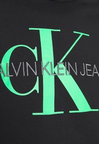 Tessuto nero con grande testo verde e grigio che scrive "ALVIN KLEIN JEANS," con un accentuato "CK" in verde brillante.