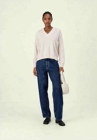Un pull rose clair à col en V avec des manches longues, assorti à un jean en denim bleu foncé ample. Le modèle tient un petit sac de couleur claire.