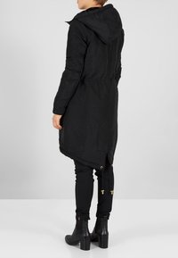 Černá dlouhá bunda s kapucí, elastickým pasem a detaily na zadním zipu, vyrobená z hladké, matné látky. Má zúžený střih a stahovací šňůrku.
